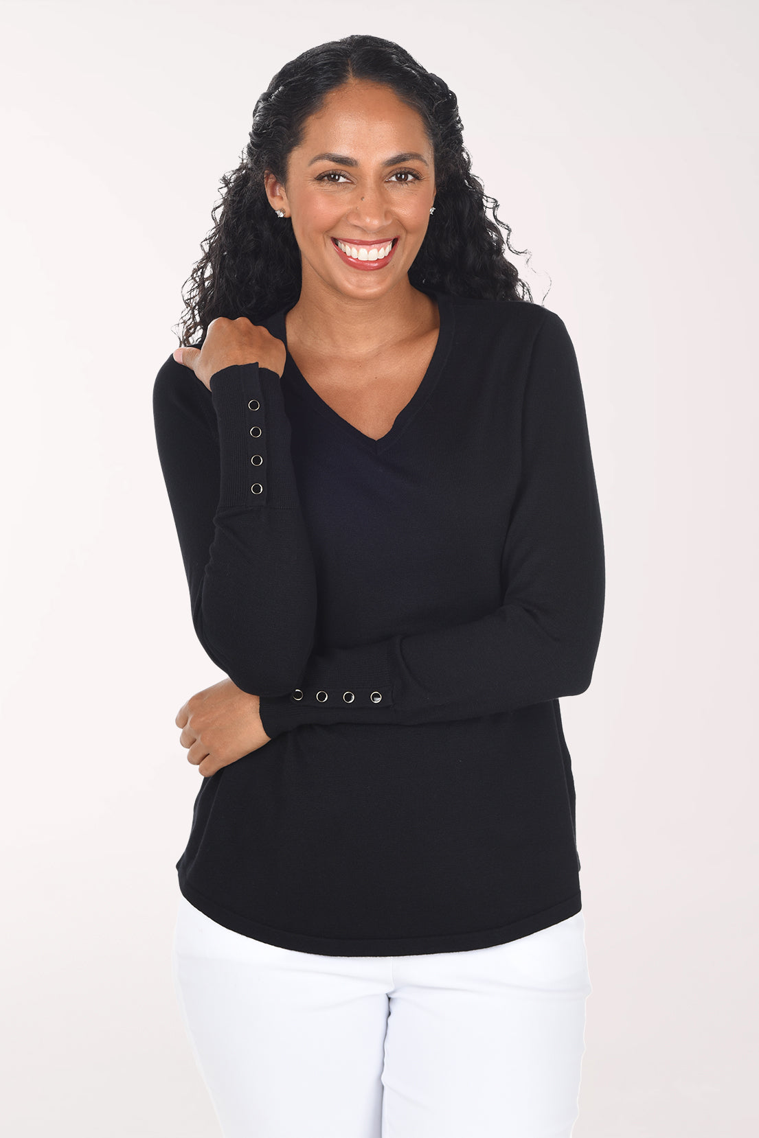 A smiling woman with long curly black hair wearing a black long-sleeve V-neck top with decorative buttons on the cuff and white pants, standing against a plain light background.