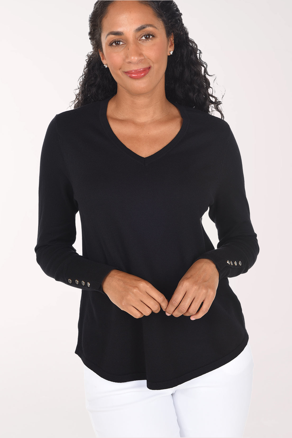 A smiling woman with long curly black hair wearing a black long-sleeve V-neck top with decorative buttons on the cuff and white pants, standing against a plain light background.