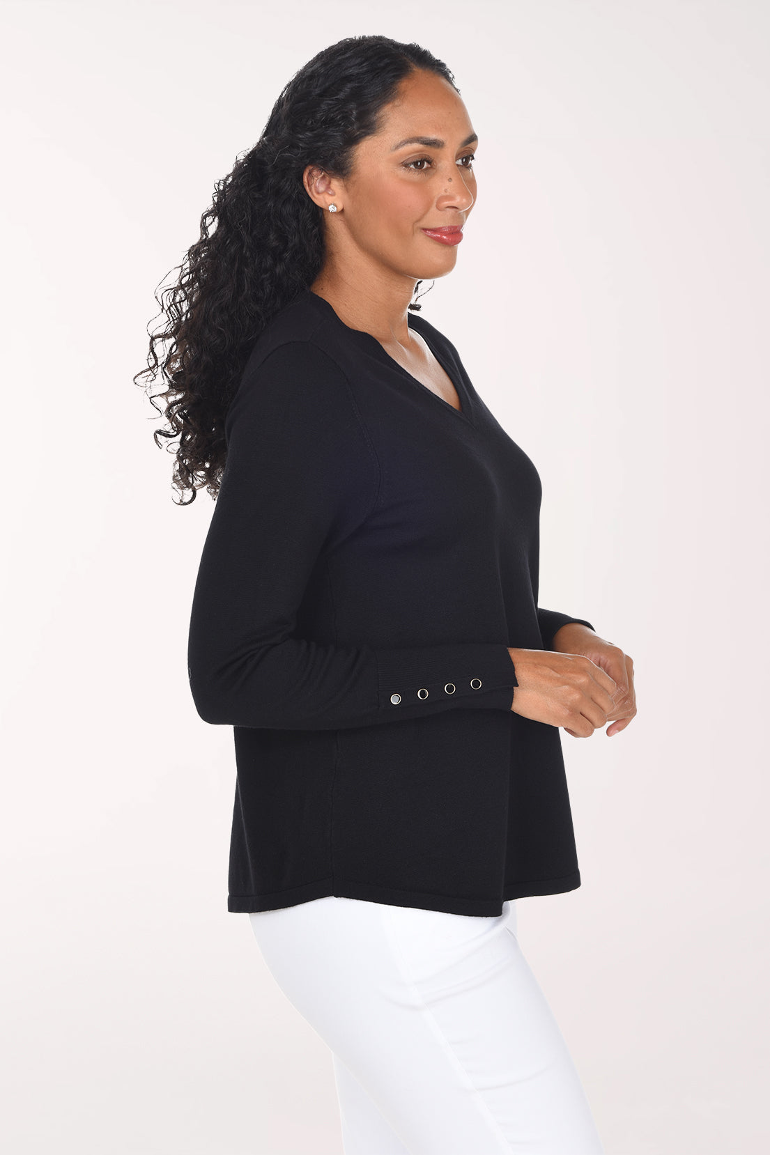 A smiling woman with long curly black hair wearing a black long-sleeve V-neck top with decorative buttons on the cuff and white pants, standing against a plain light background.