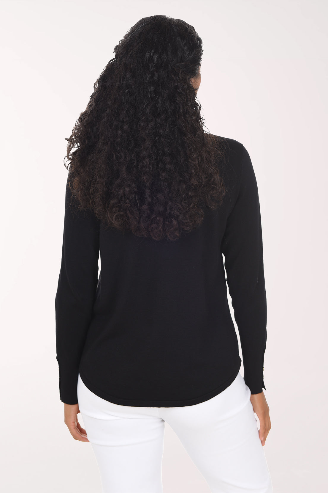 A smiling woman with long curly black hair wearing a black long-sleeve V-neck top with decorative buttons on the cuff and white pants, standing against a plain light background.