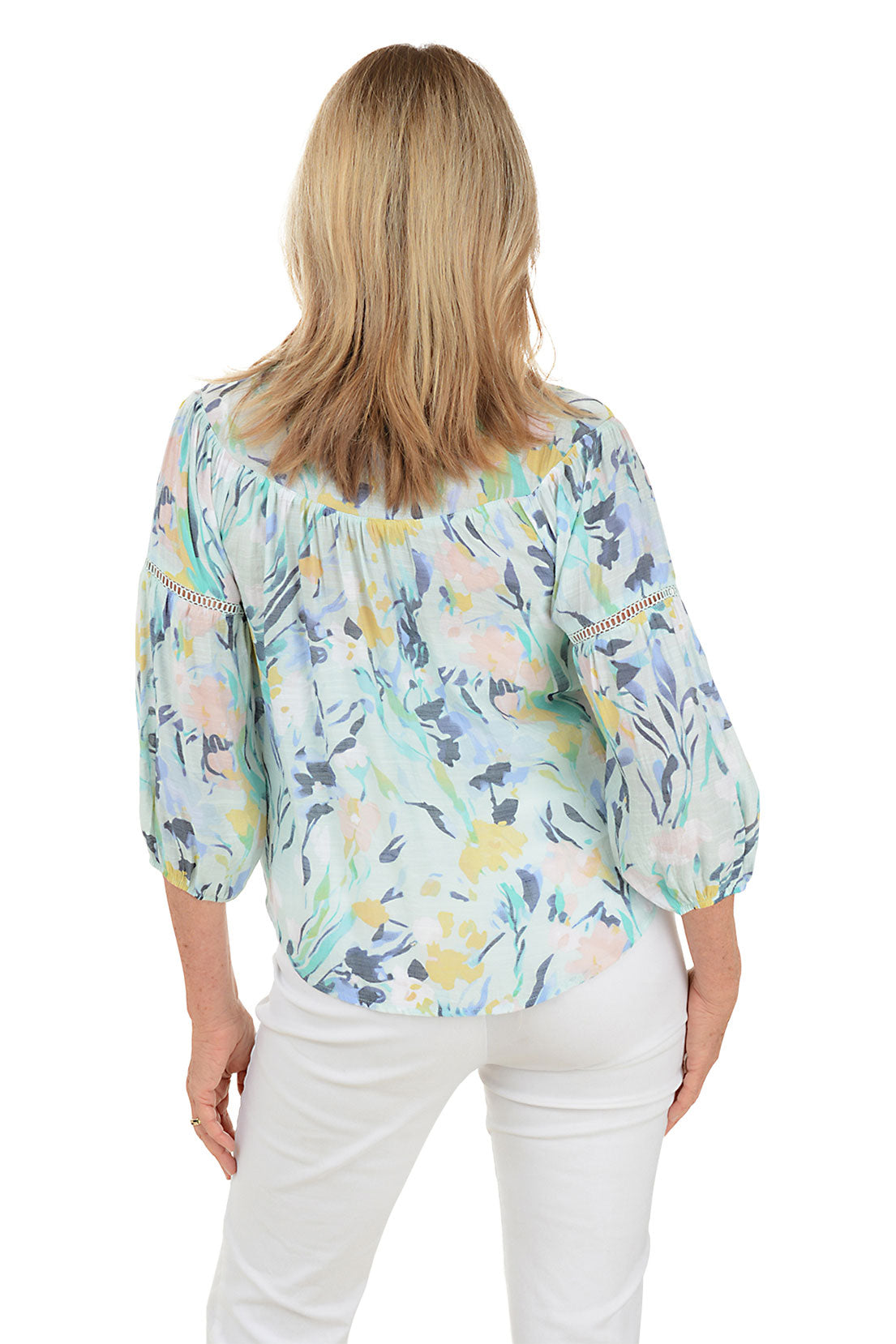 A back shot of a model wearing a floral print crochet puff sleeve blouse with a scoop neckline and buttoned half placket, paired with white pants.