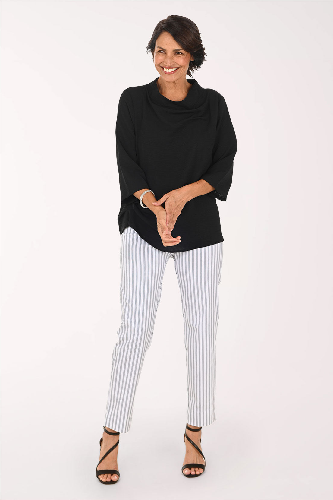 A woman modeling a black cowl-neck top and white pants with thin blue vertical stripes, paired with black strappy high-heeled sandals, against a plain white studio background.