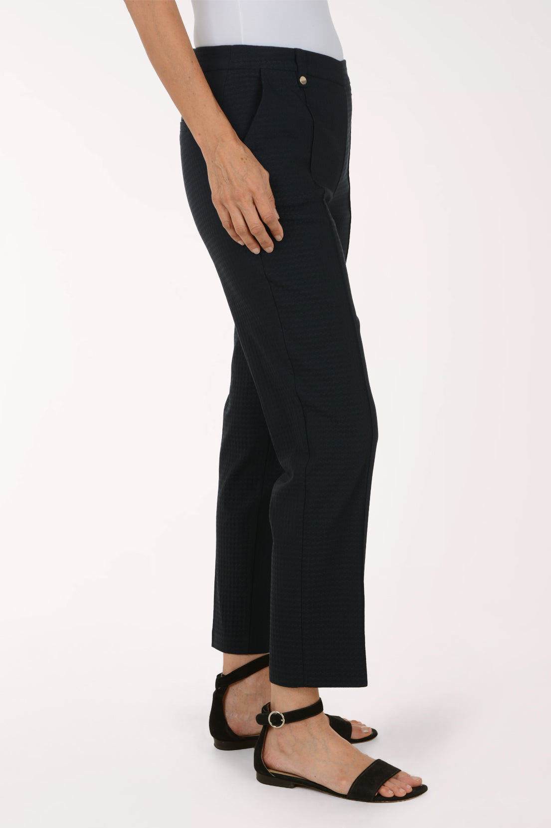 A model wearing black cropped trousers with a subtle vertical textured stripe pattern, paired with black flat sandals and a white top, shown from the waist down against a plain white studio background.