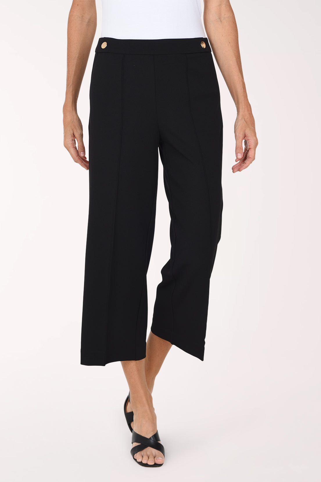 A model wearing black cropped wide-leg trousers with gold button details at the waistband, paired with a white top and black sandals, against a plain light background.