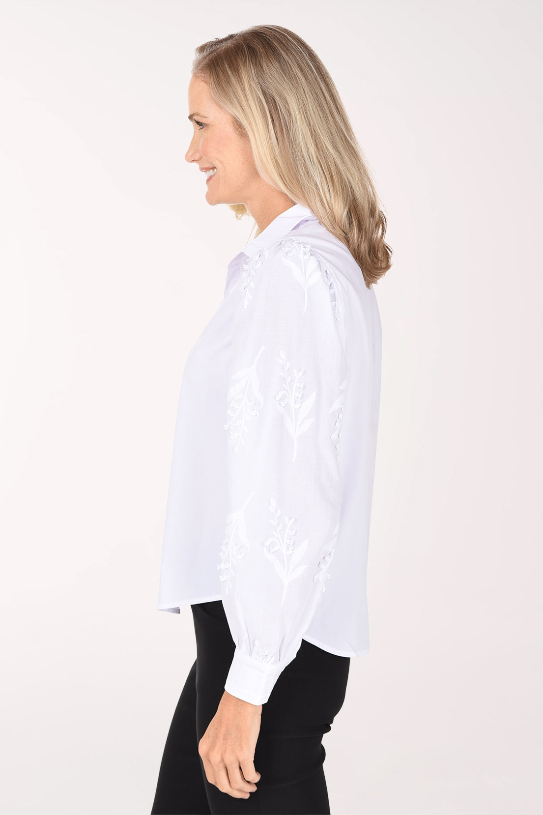 A woman wearing a white long-sleeve blouse with delicate floral embroidery on the sleeves and black pants, photographed against a plain white studio background.