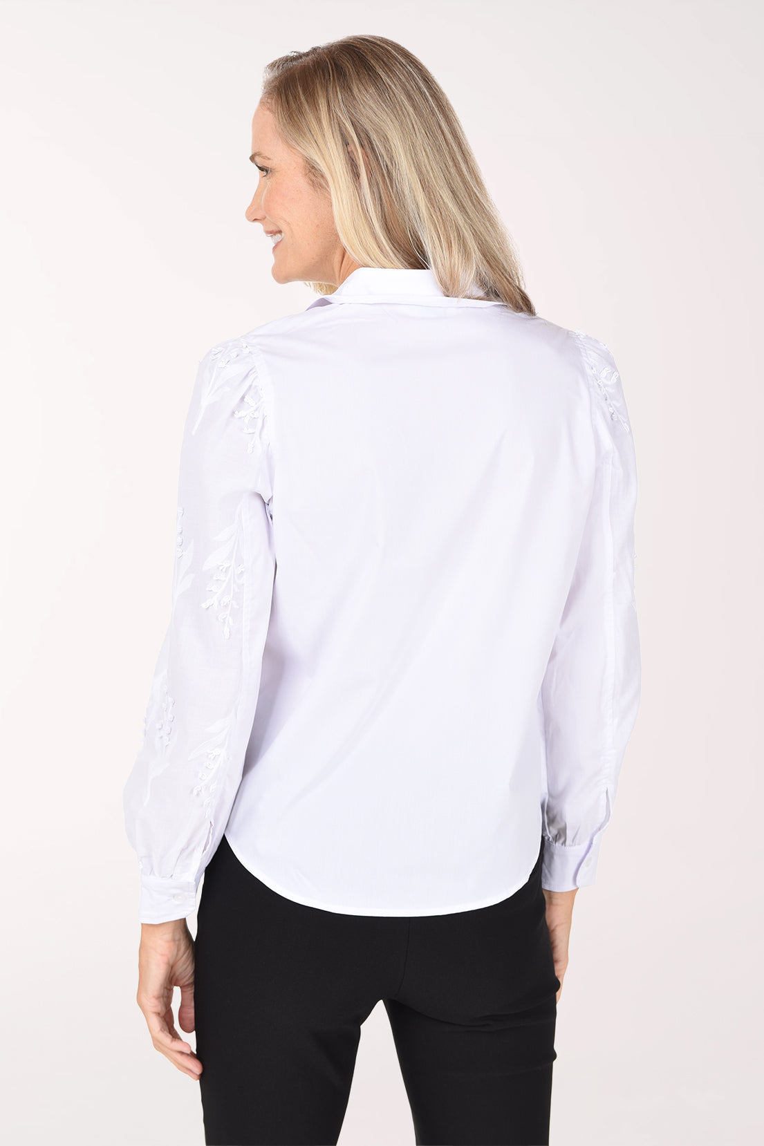 A woman wearing a white long-sleeve blouse with delicate floral embroidery on the sleeves and black pants, photographed against a plain white studio background.