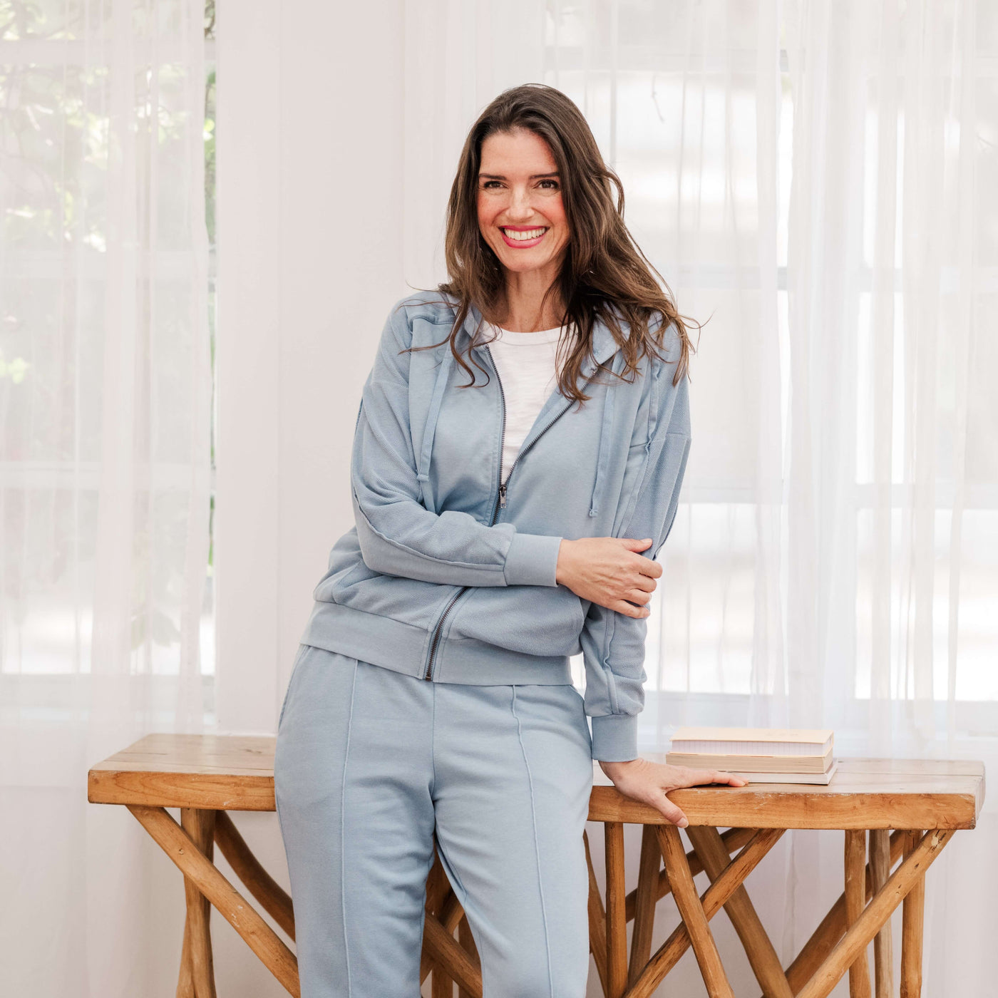 A woman in a light blue hoodie and matching pants leans against a wooden table with a curtained window behind her