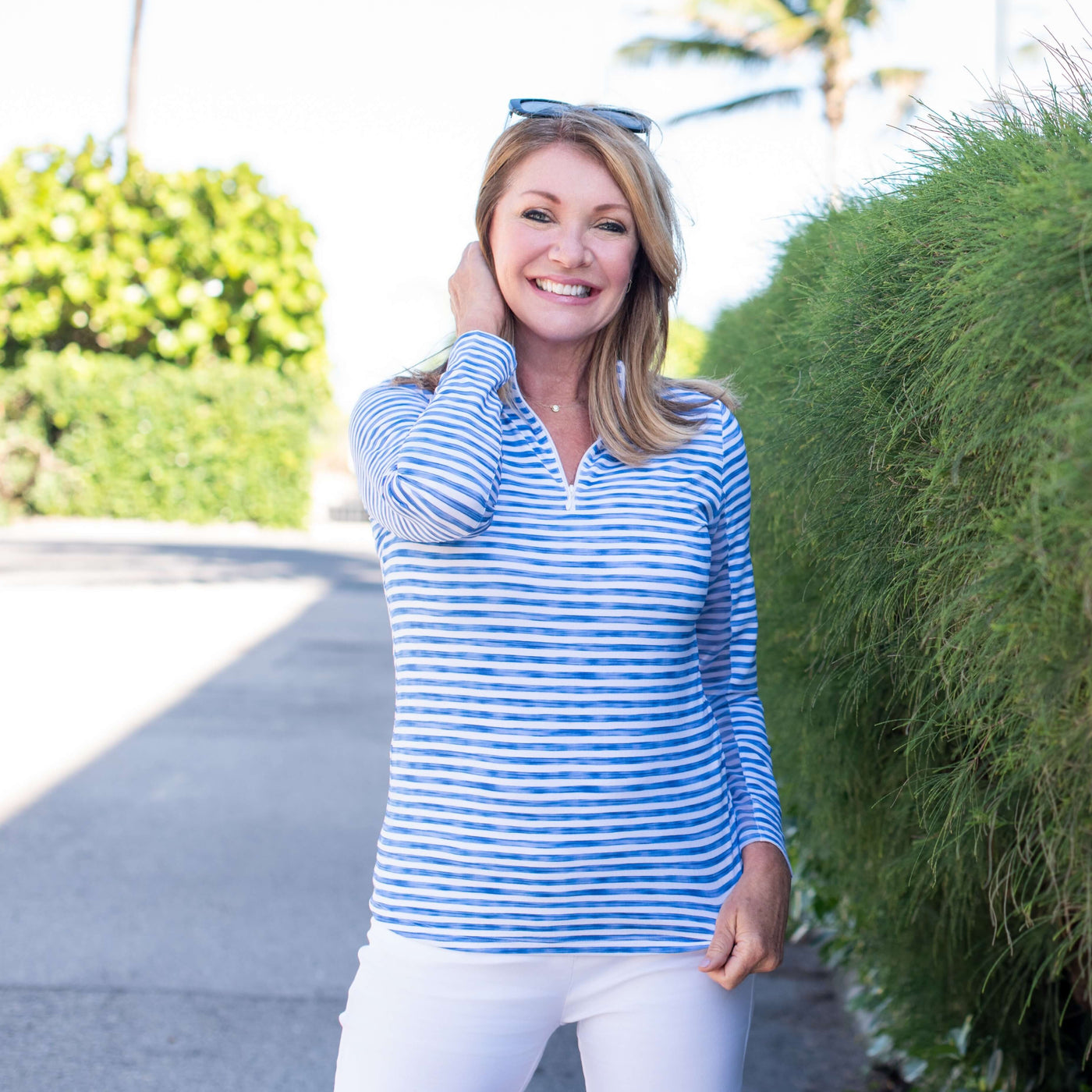 A smiling woman in a white and blue striped sun shirt and white shorts stands in the shade next to a green hedge.