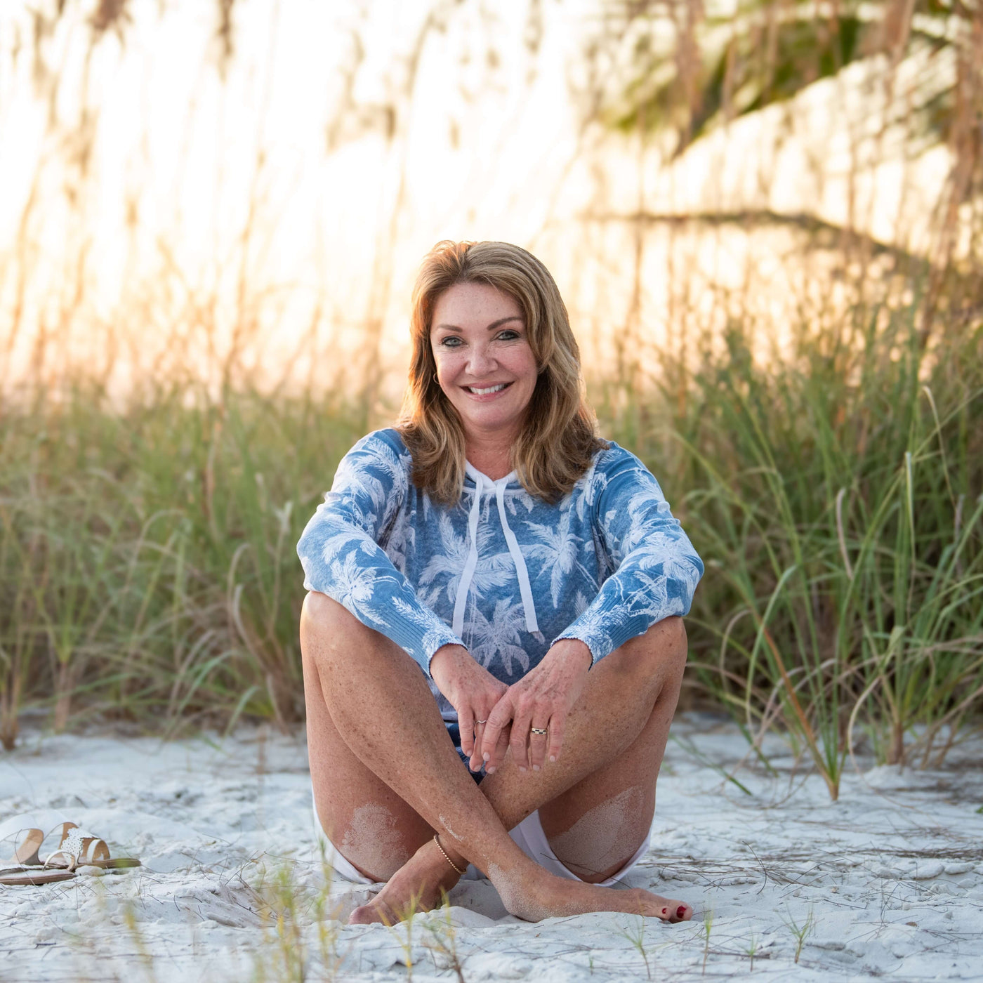 Woman sitting on a sandy beach wearing a blue and white hoodie and white shorts