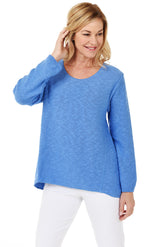 A woman wearing a long sleeve ocean blue cotton scoop neck sweater, with a seam down the front and white pants.