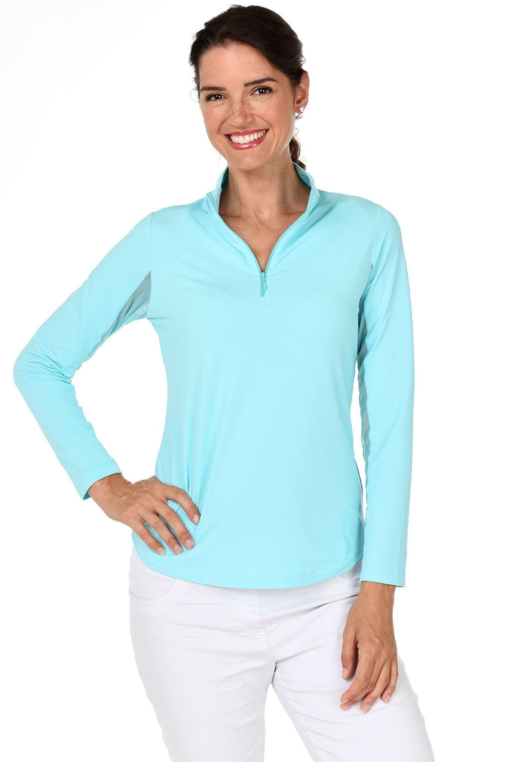 A woman smiling and wearing a long-sleeved seafoam sun shirt with a mock-neck collar and a pair of white pants.