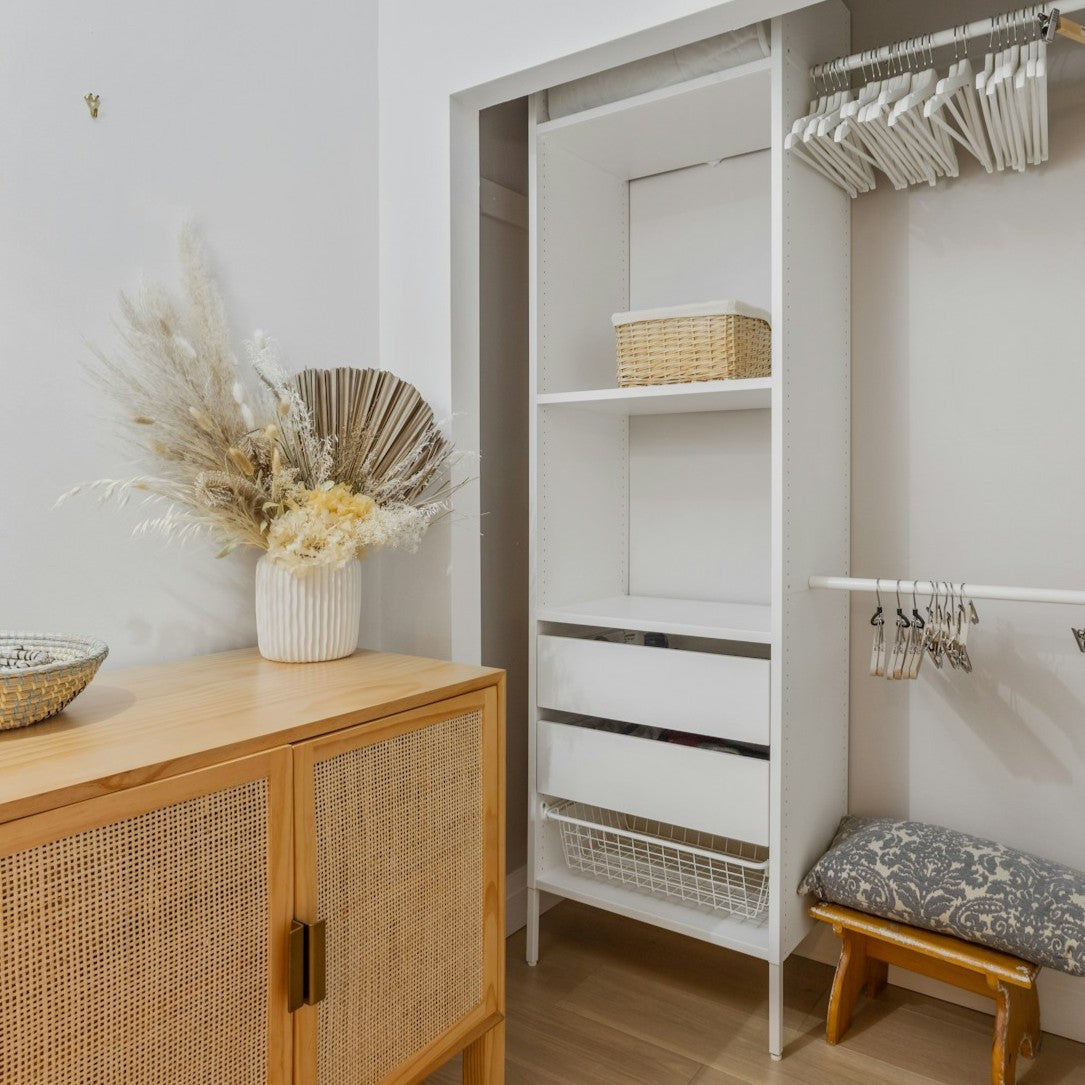 An empty closet with hangers next to a rattan cabinet with a vase of dried flowers
