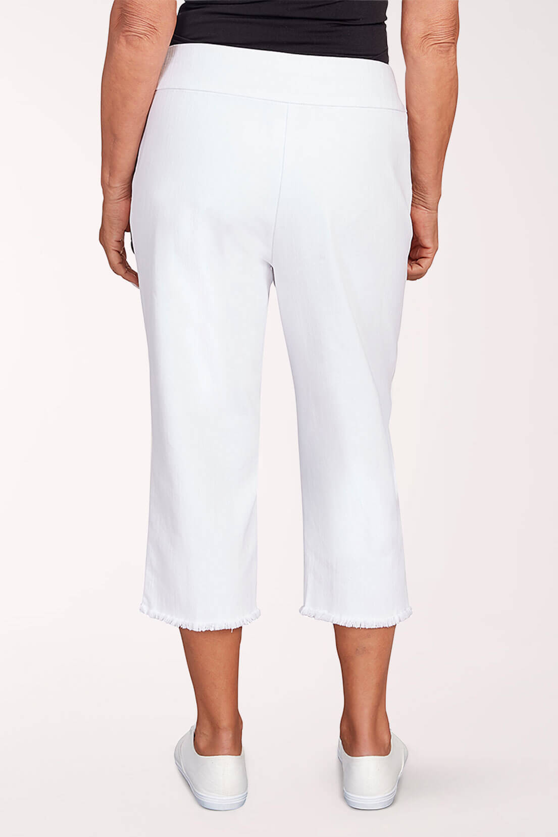 A woman wearing white denim capri pants and white sneakers against a light pink background. Back shot