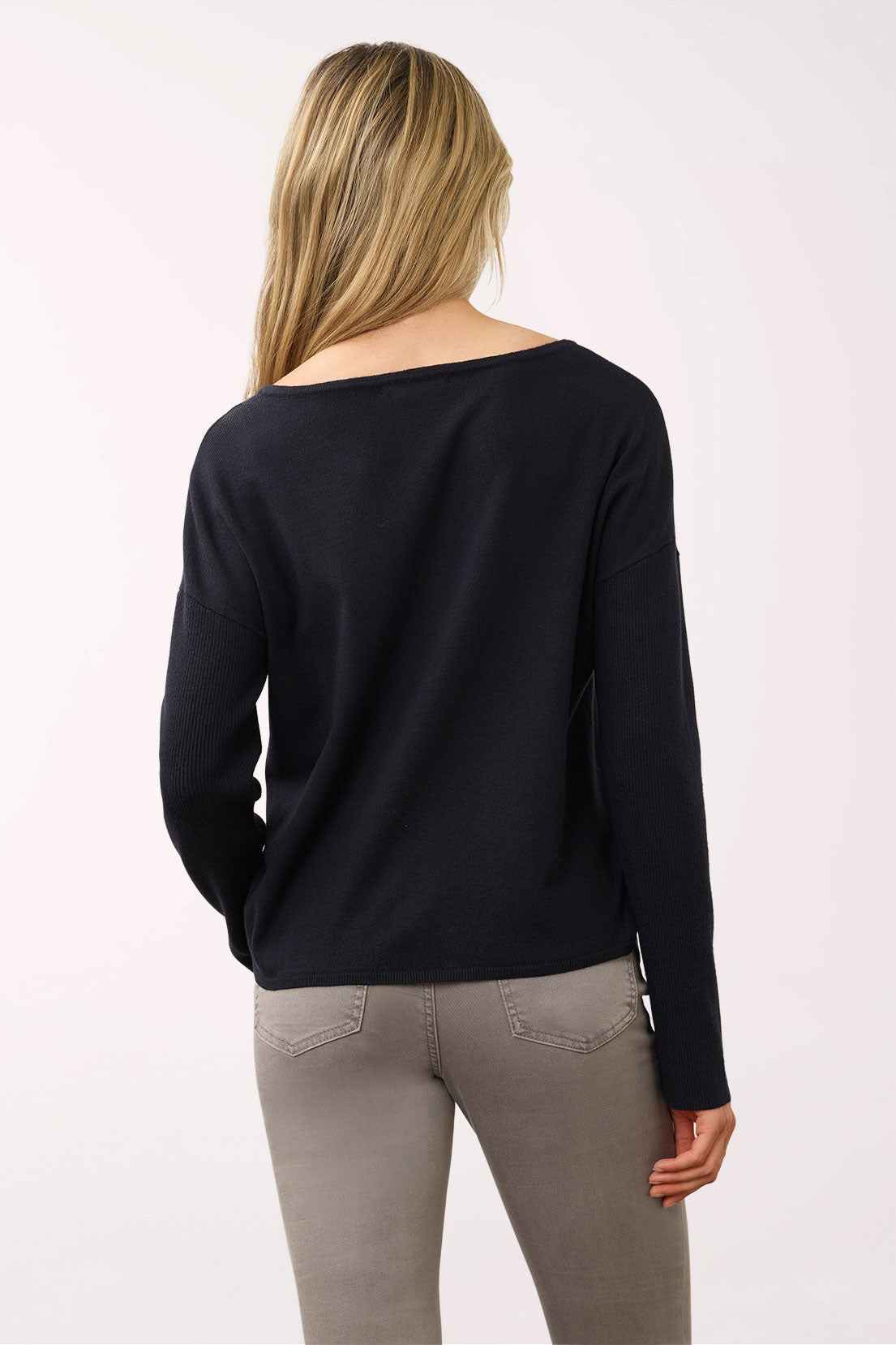 A woman viewed from the back wearing a black long-sleeve boat-neck top and gray pants, photographed against a plain light background.