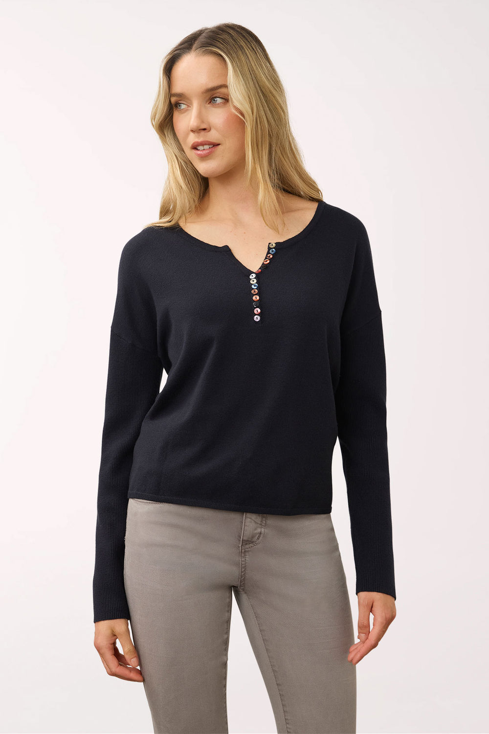 A woman modeling a black long-sleeve knit top with a decorative buttoned split neckline, paired with gray pants, against a plain light background.
