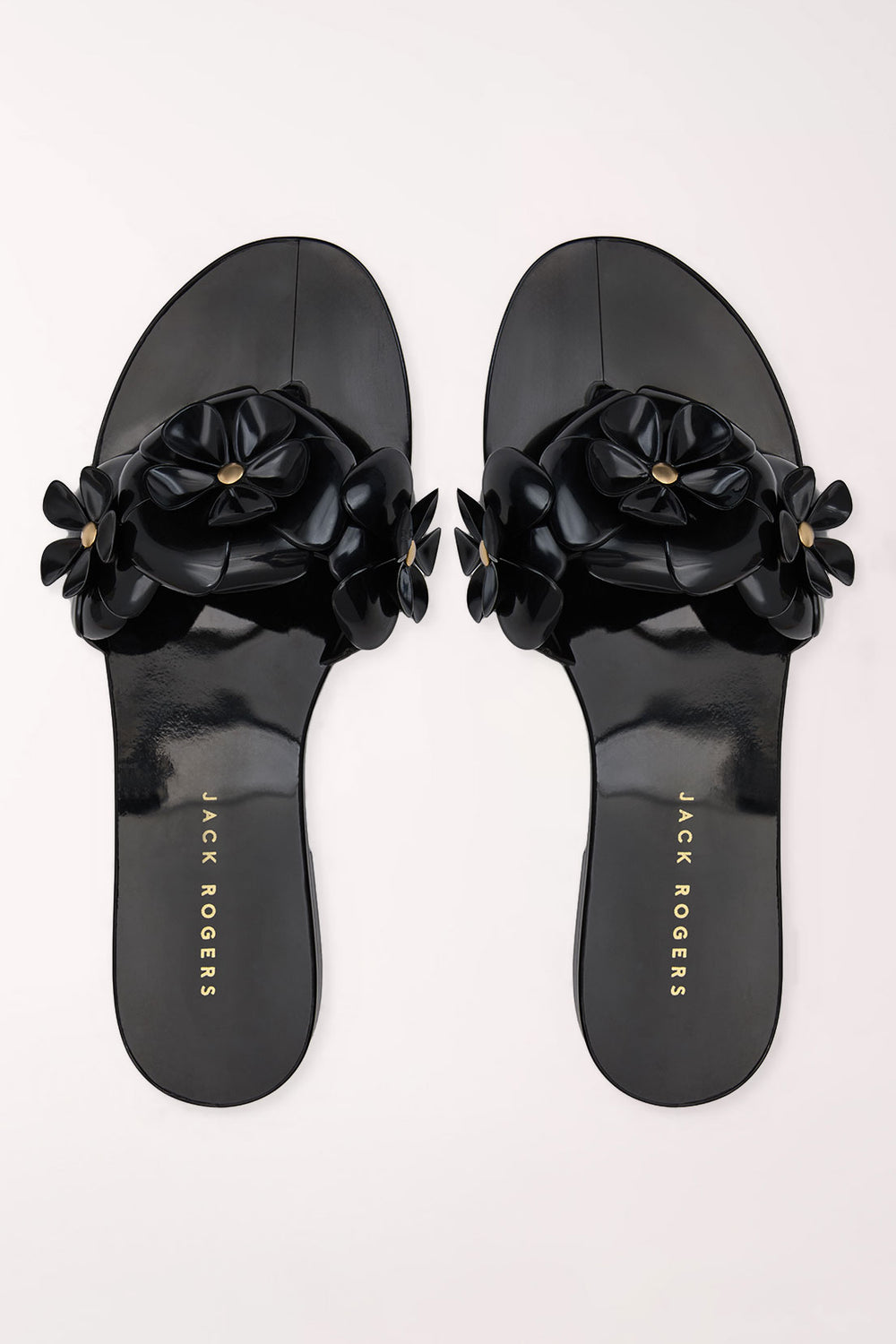 A pair of black patent jelly slide sandals by Jack Rogers, featuring large 3D floral embellishments across the straps with small gold-tone center details, shown from a top-down view on a light off-white background.