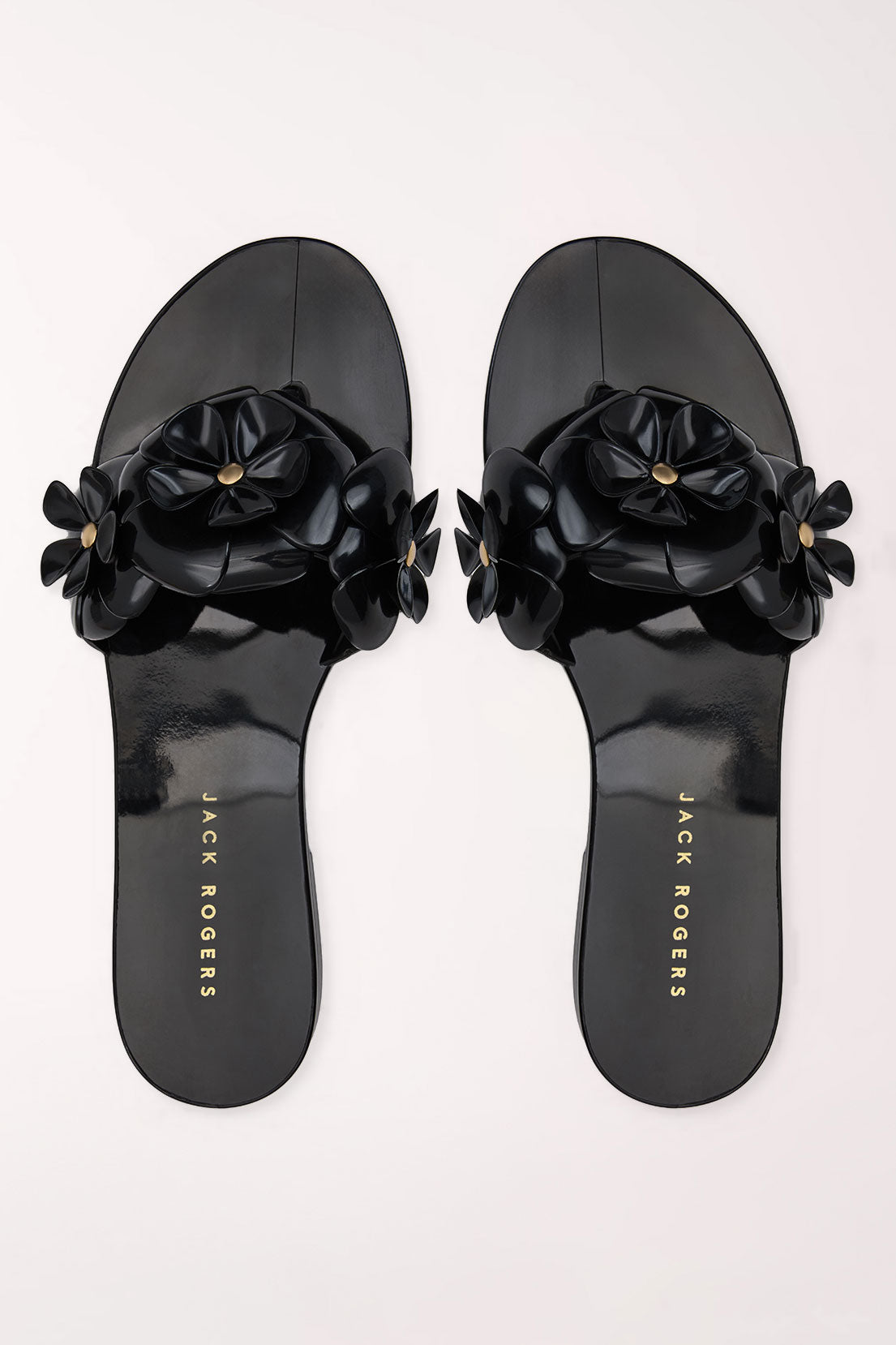 A pair of black patent jelly slide sandals by Jack Rogers, featuring large 3D floral embellishments across the straps with small gold-tone center details, shown from a top-down view on a light off-white background.