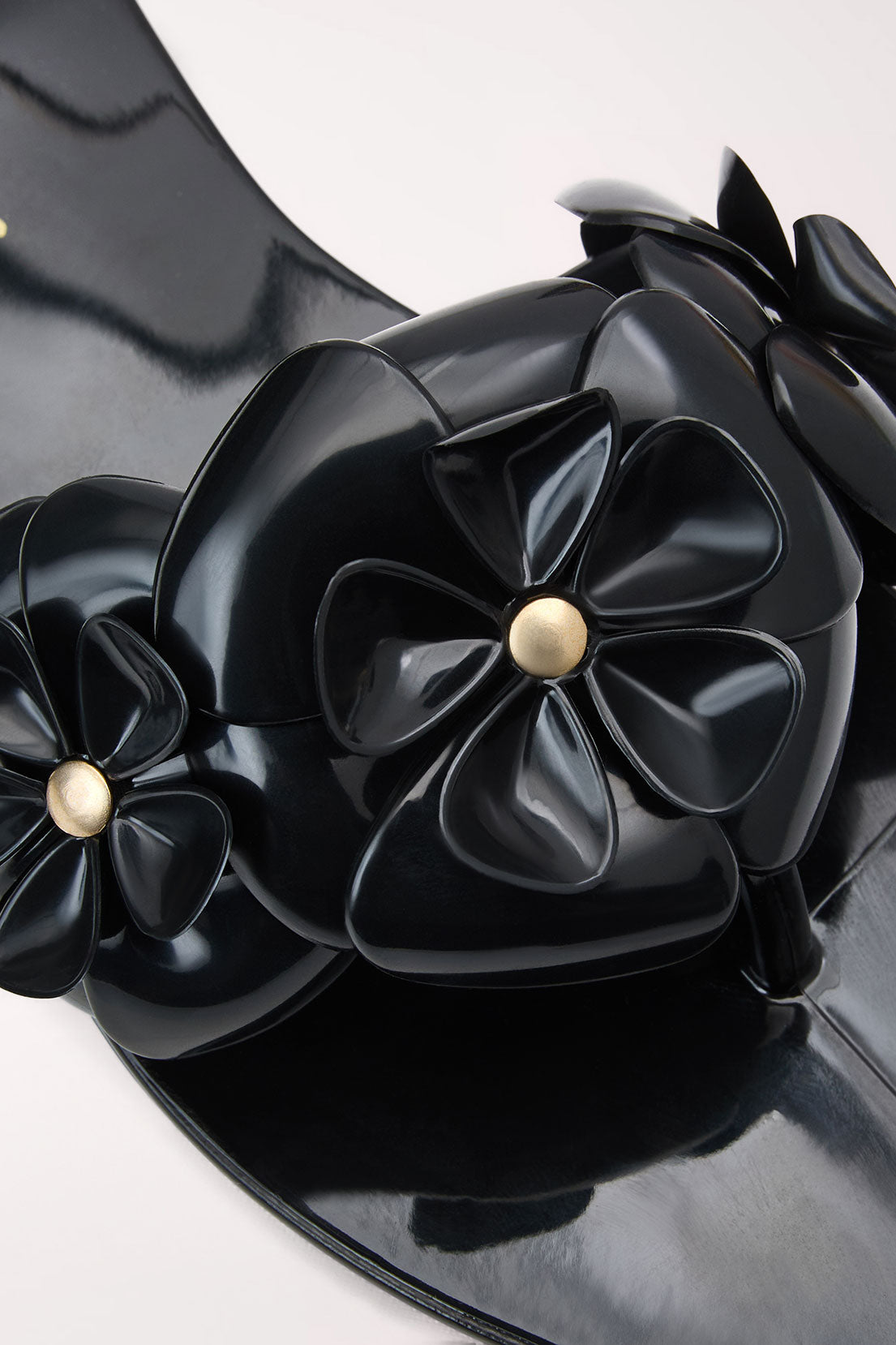 Close-up of a black patent leather accessory with large sculptural flower appliqués and small gold-tone center studs on a light gray background.