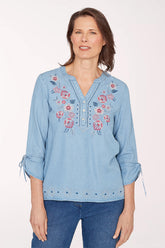 Woman wearing a light blue denim blouse with floral embroidery on a white background