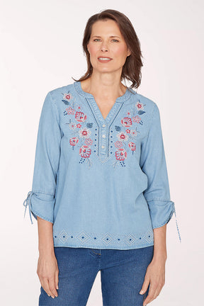 Woman wearing a light blue denim blouse with floral embroidery on a white background
