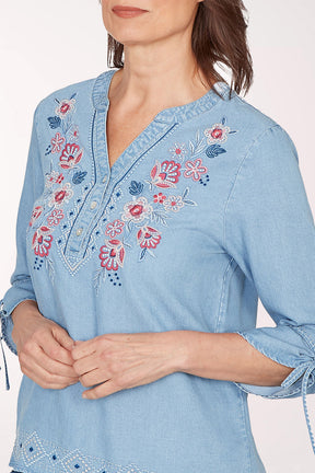 Woman wearing a light blue denim blouse with floral embroidery on a white background