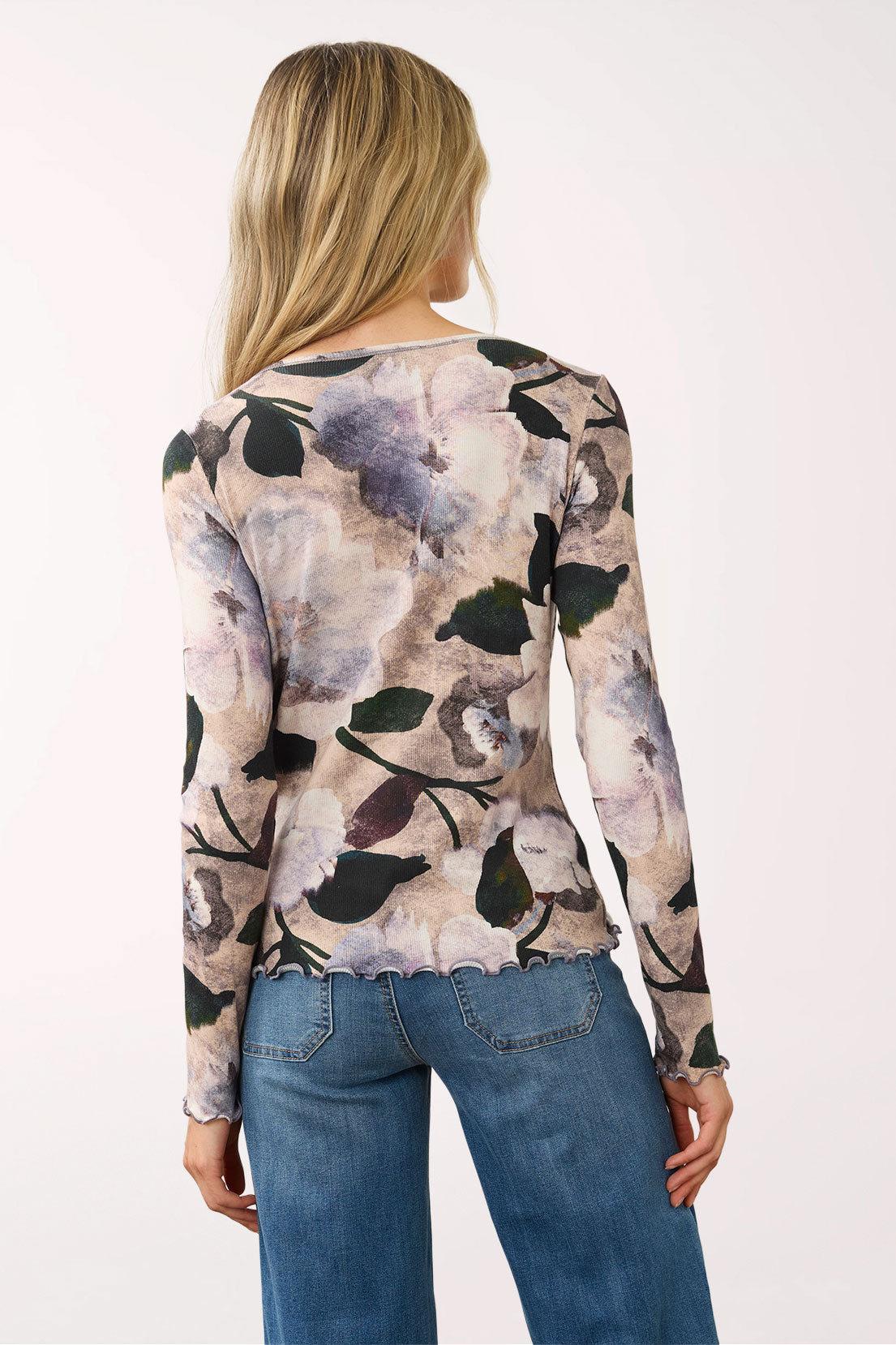 A woman viewed from the back wearing a long-sleeve knit top with a muted floral and abstract print in beige, gray, and black, paired with blue jeans against a plain light background.