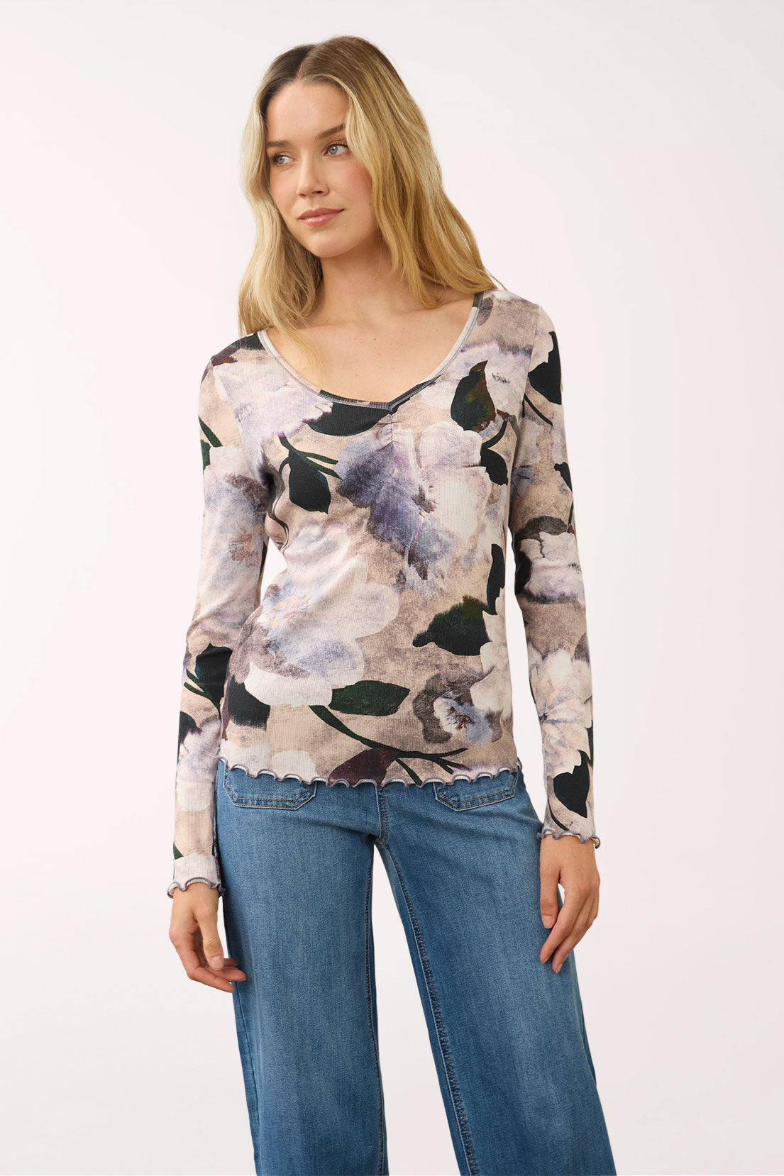 A woman modeling a long-sleeve scoop-neck top with a muted floral print in beige, gray, black, and lavender, paired with blue jeans against a plain white studio background.