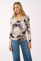 A woman modeling a long-sleeve scoop-neck top with a muted floral print in beige, gray, black, and lavender, paired with blue jeans against a plain white studio background.