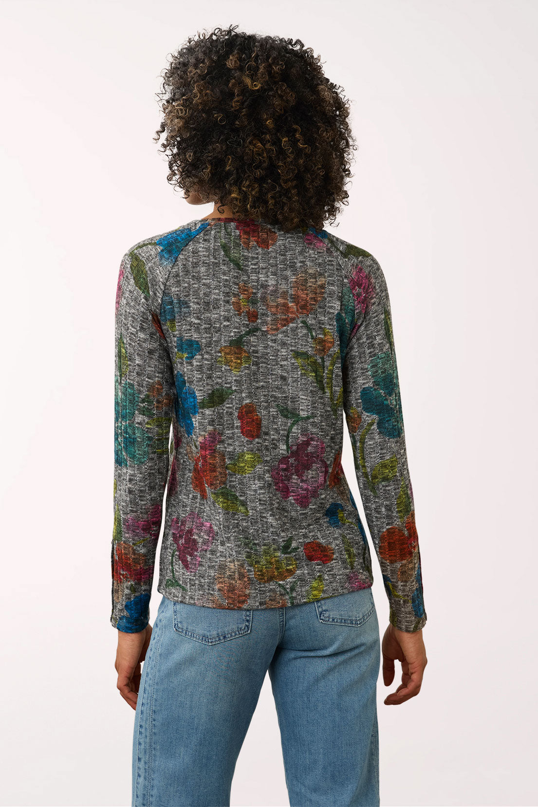 A woman viewed from behind wearing a long-sleeve knit top with a textured gray base and a colorful floral pattern, paired with light blue jeans against a plain white background.