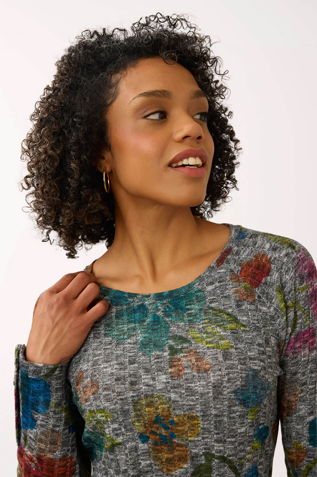 A woman with curly dark hair wearing a long-sleeve gray knit top with a multicolor floral pattern, photographed against a plain light background.