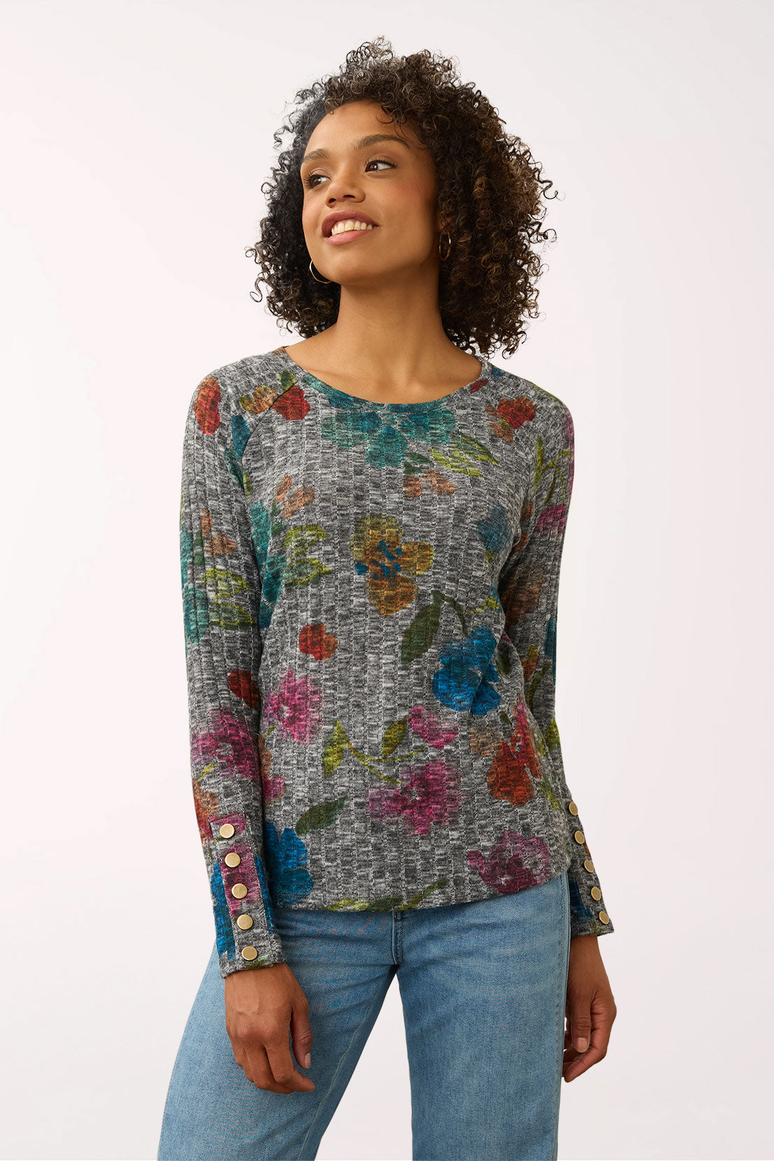 A woman wearing a long-sleeve knit top with a multicolor floral pattern on a gray textured background, paired with blue jeans, photographed against a plain light background.
