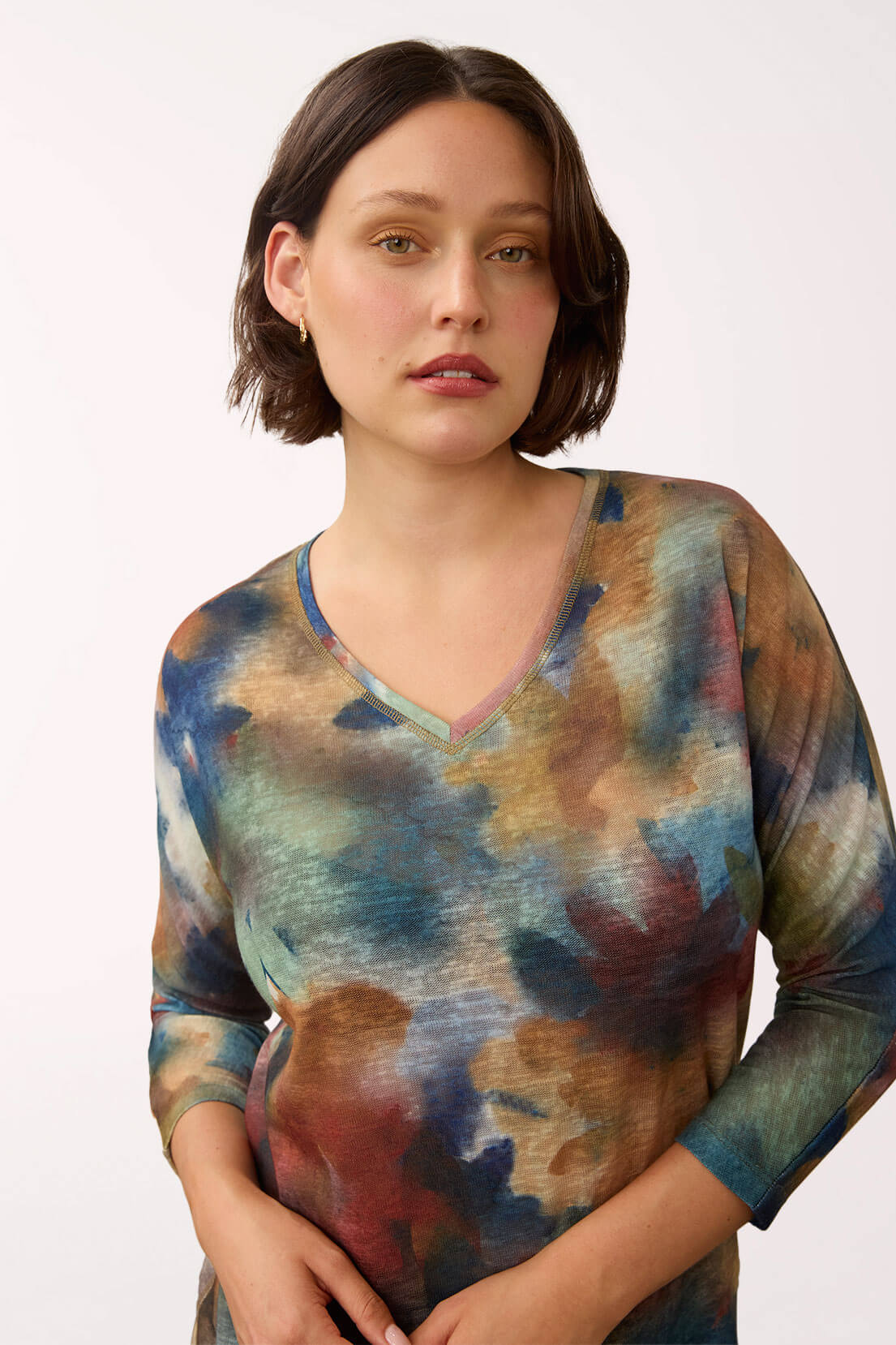 A woman wearing a three-quarter sleeve V-neck top with an abstract watercolor-like print in shades of blue, teal, brown, and rust, photographed against a plain light background.