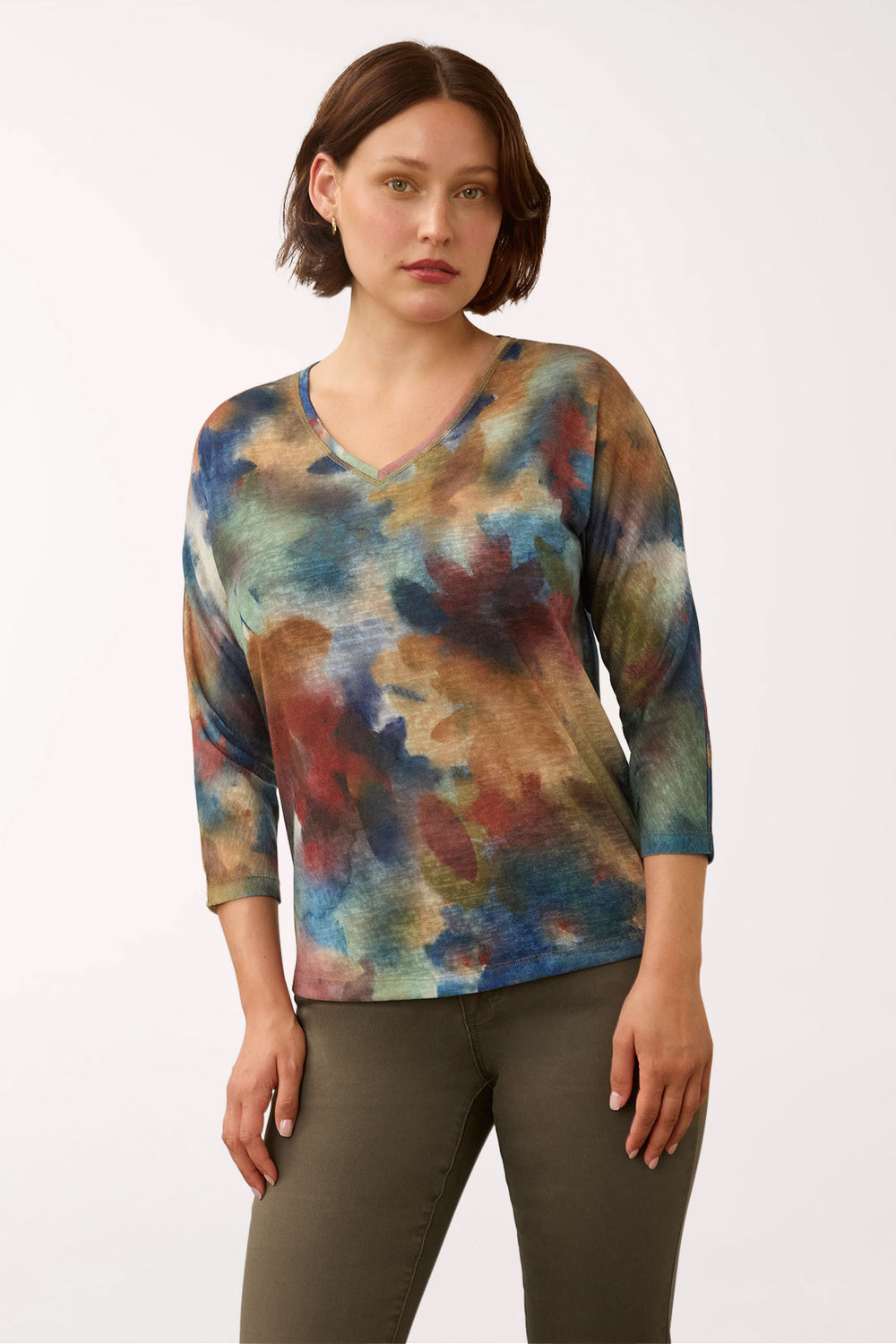 A woman modeling a three-quarter sleeve V-neck top with an abstract watercolor print in shades of blue, brown, red, and beige, paired with olive green pants against a plain white studio background.