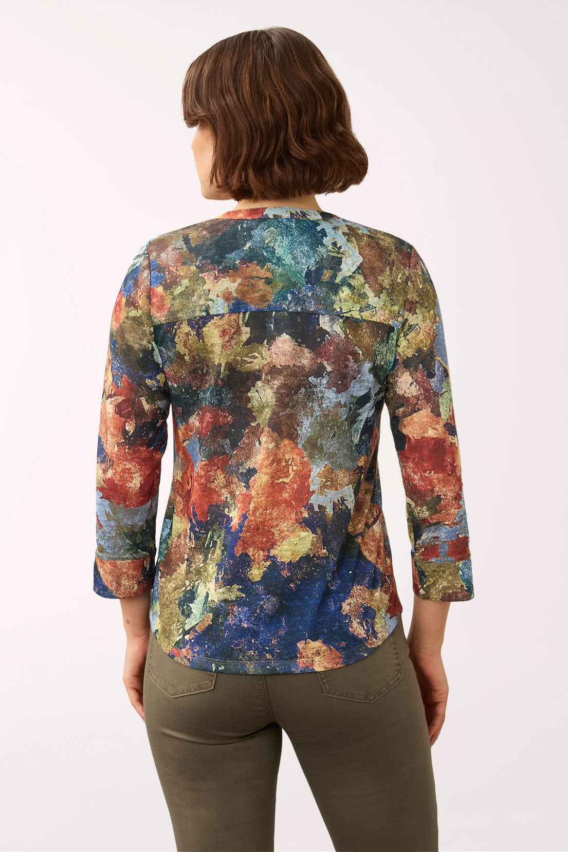 A woman viewed from behind wearing a long-sleeve button-up blouse with a vibrant, multicolored abstract floral print and olive green pants, set against a plain white studio background.