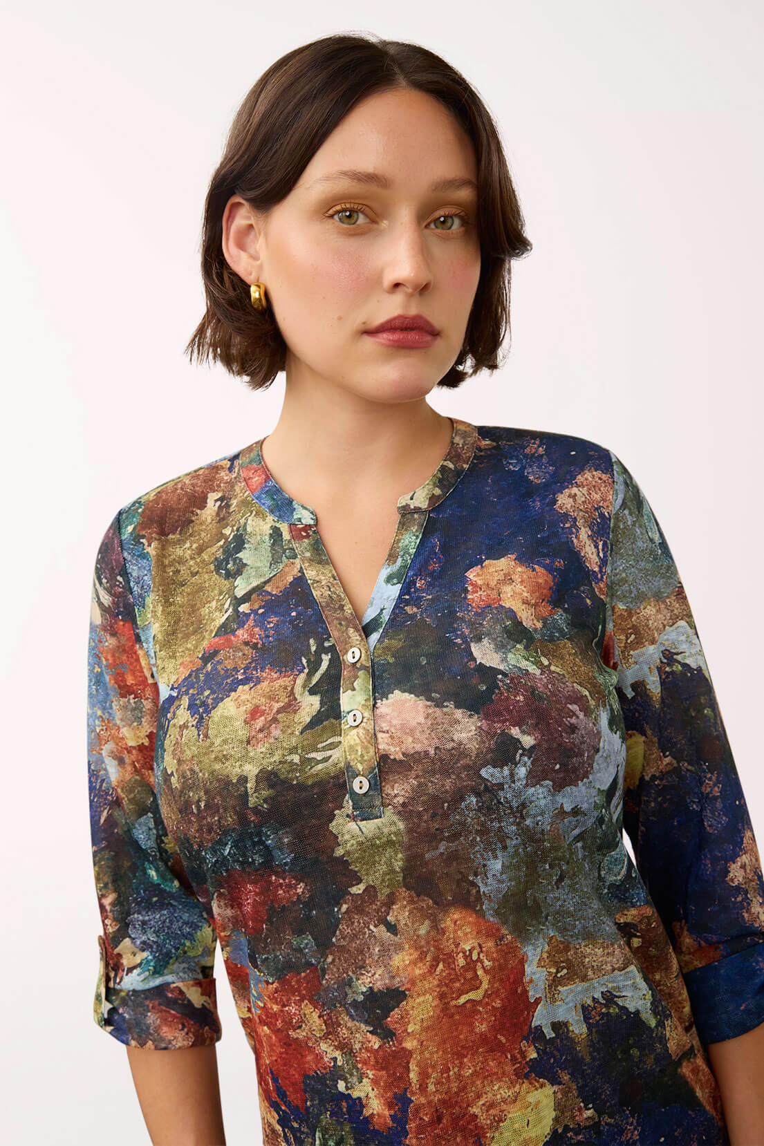 A woman wearing a long-sleeve button-front top with a multicolor abstract painterly print in blue, red, brown, and beige tones, photographed against a plain light background.