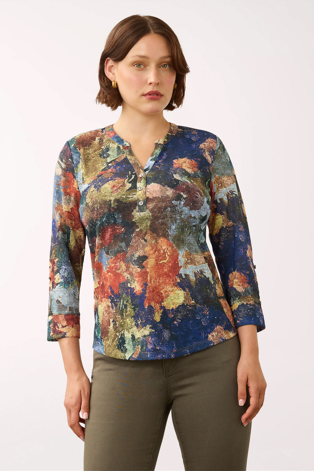 A woman modeling a long-sleeve V-neck blouse with an abstract painterly print in blue, red, orange, yellow, and green, paired with olive green pants against a plain white studio background.