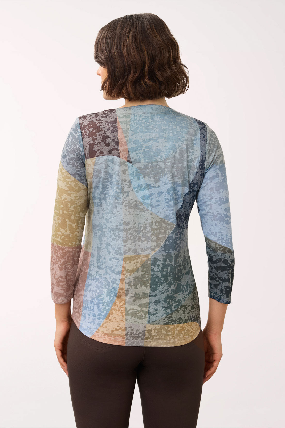 A woman viewed from the back wearing a long-sleeve patchwork top with an abstract, distressed print in shades of blue, beige, brown, and gray, paired with dark brown leggings, against a plain white background.
