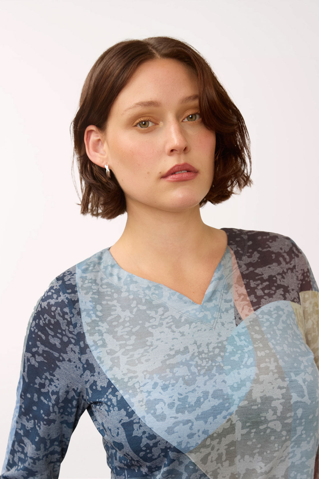 A woman with a short brown bob haircut wearing a V-neck long-sleeve top with a blue, gray, and beige abstract pattern, photographed against a plain light background.