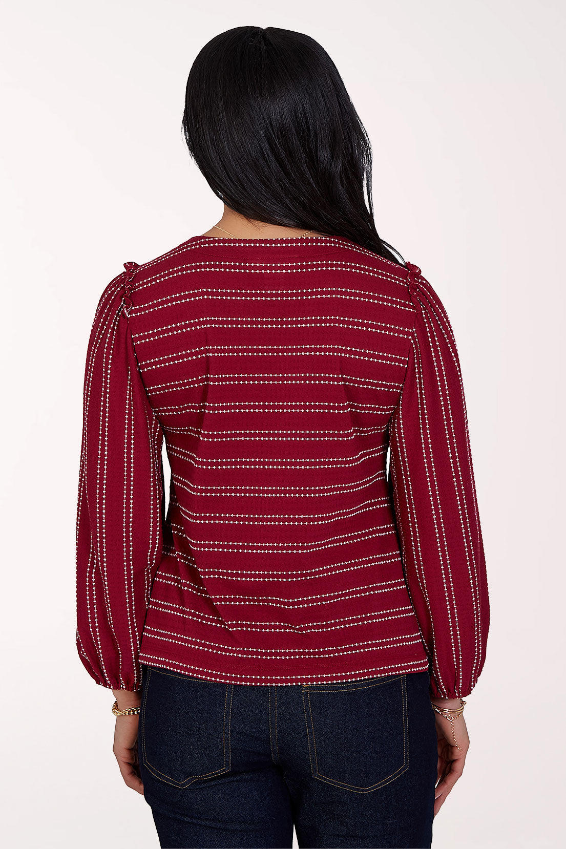 A woman viewed from the back wearing a burgundy long-sleeve top with thin white horizontal stripes and dark blue jeans, set against a plain off-white background.