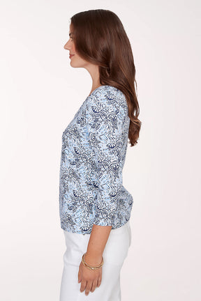 Woman wearing a blue patterned top with white pants on a plain background