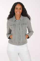 A woman with long curly dark hair wearing a light gray washed denim jacket and white pants, posing against a solid white studio background.