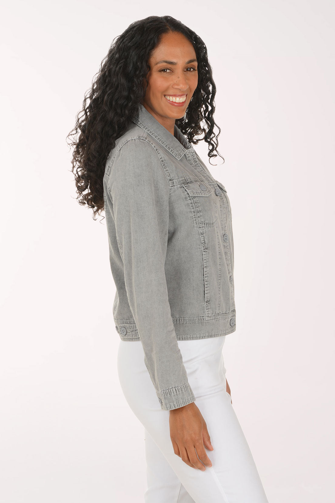 A woman with long curly dark hair wearing a light gray washed denim jacket and white pants, posing against a solid white studio background.