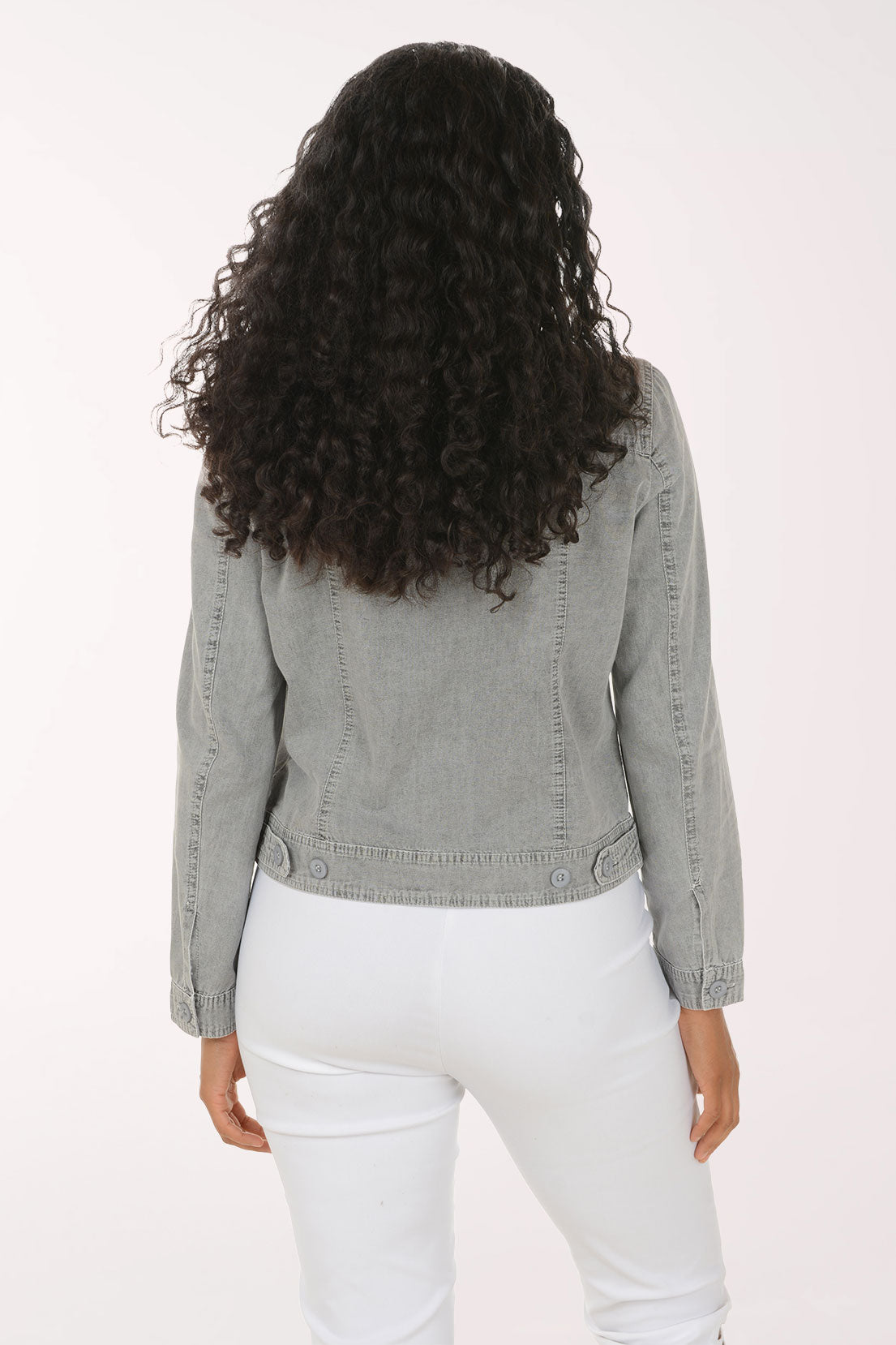 A woman with long curly dark hair wearing a light gray washed denim jacket and white pants, posing against a solid white studio background.
