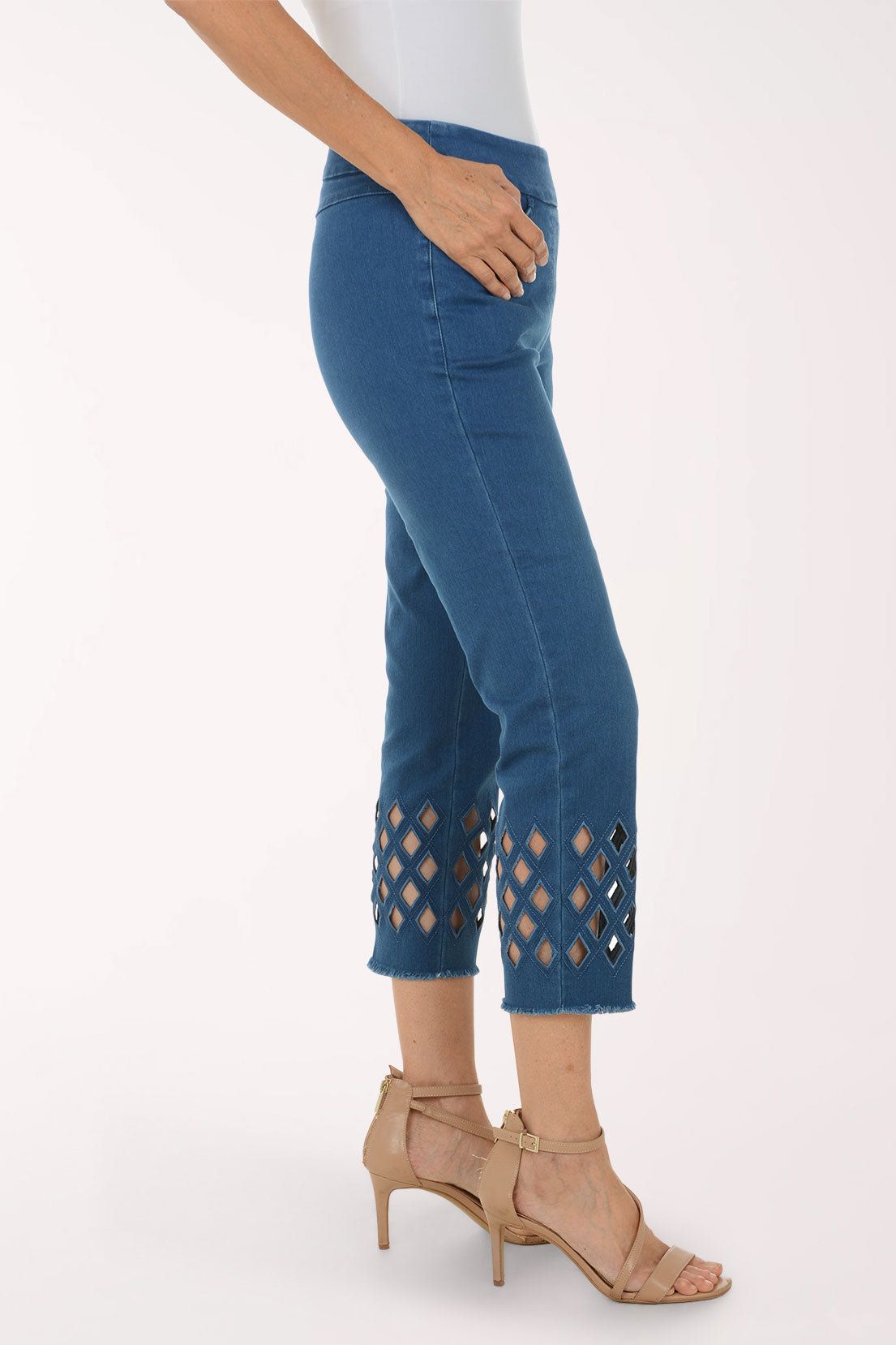 A model wearing blue cropped jeans with diamond-shaped cutout details near the hem, paired with a white top and beige strappy high-heeled sandals, against a plain white studio background.