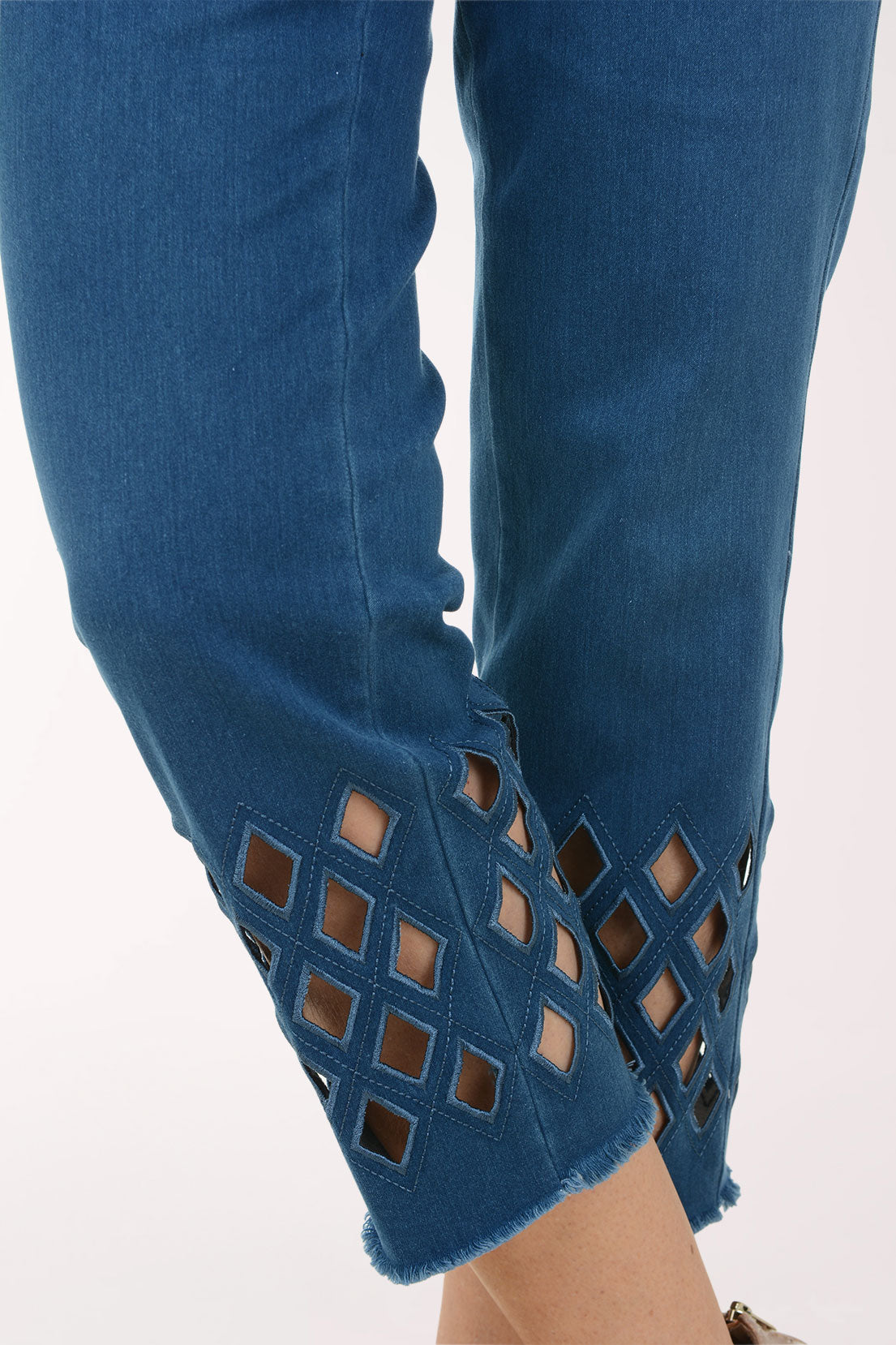 A model wearing blue cropped jeans with diamond-shaped cutout details near the hem, paired with a white top and beige strappy high-heeled sandals, against a plain white studio background.