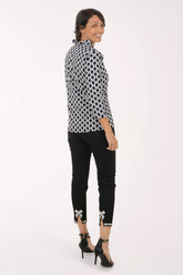 A woman modeling a black and white geometric patterned long-sleeve blouse with black cropped pants featuring bow details at the cuffs, and black high heels, photographed against a plain white studio background.