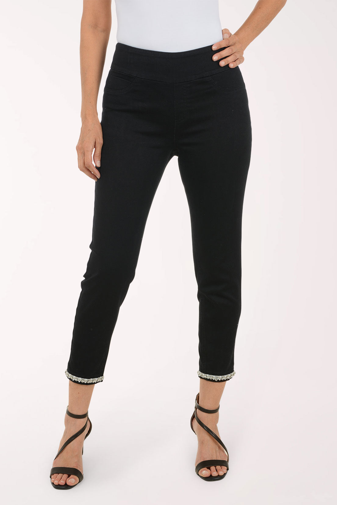 A model wearing black cropped ankle pants with a subtle beaded trim at the hem, paired with a white top and black strappy heels, against a plain white studio background.