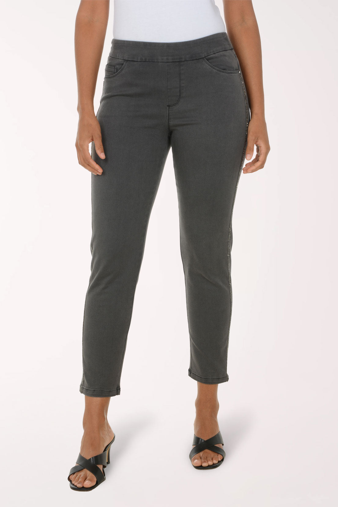 A model wearing a pair of dark gray slim-fit cropped jeans with a straight leg, paired with a white top and black heeled sandals, against a plain white studio background.