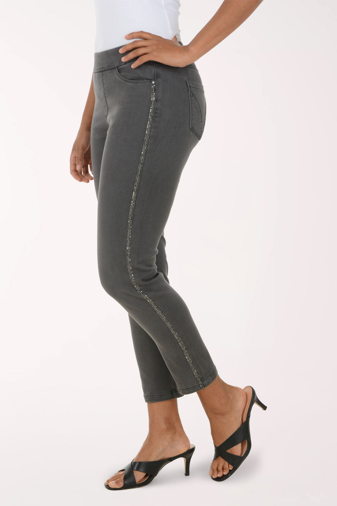 A model wearing a pair of dark gray slim-fit cropped jeans with a straight leg, paired with a white top and black heeled sandals, against a plain white studio background.