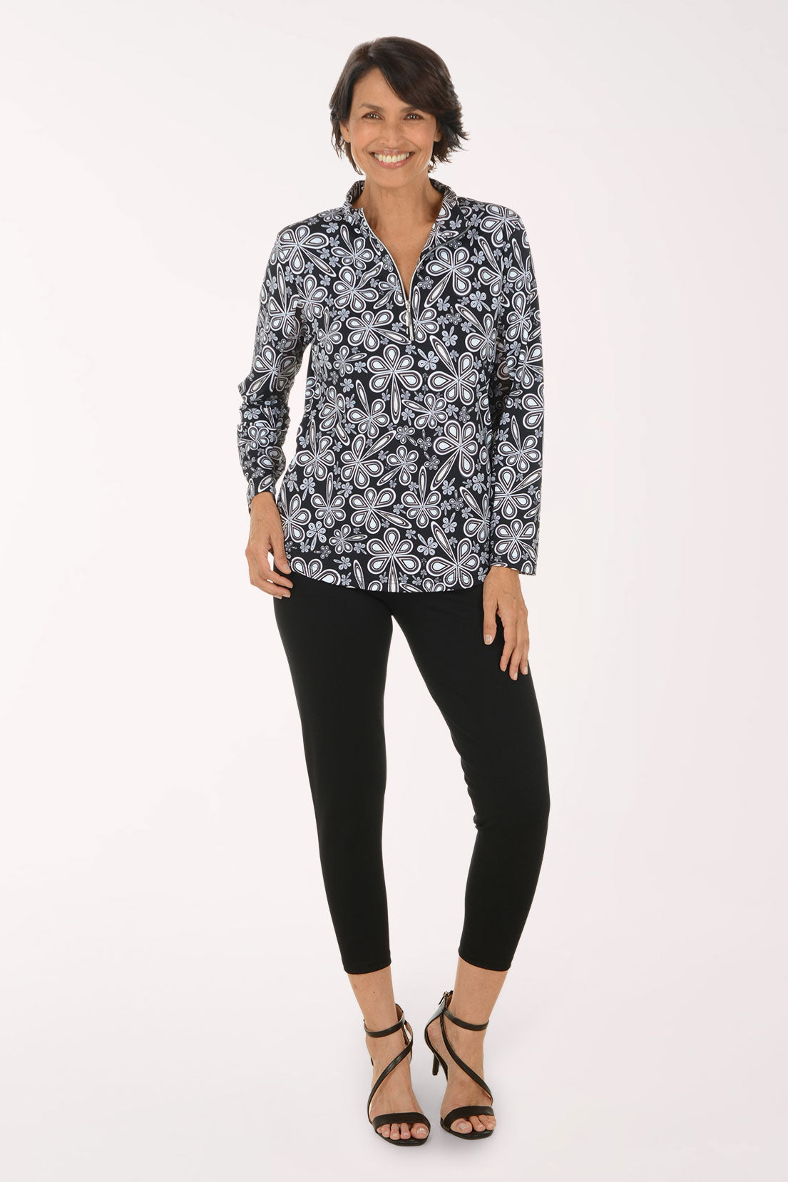 A woman modeling a long-sleeve button-up blouse with a black, white, and gray paisley pattern, paired with black cropped pants and black strappy high-heeled sandals, against a plain white studio background.
